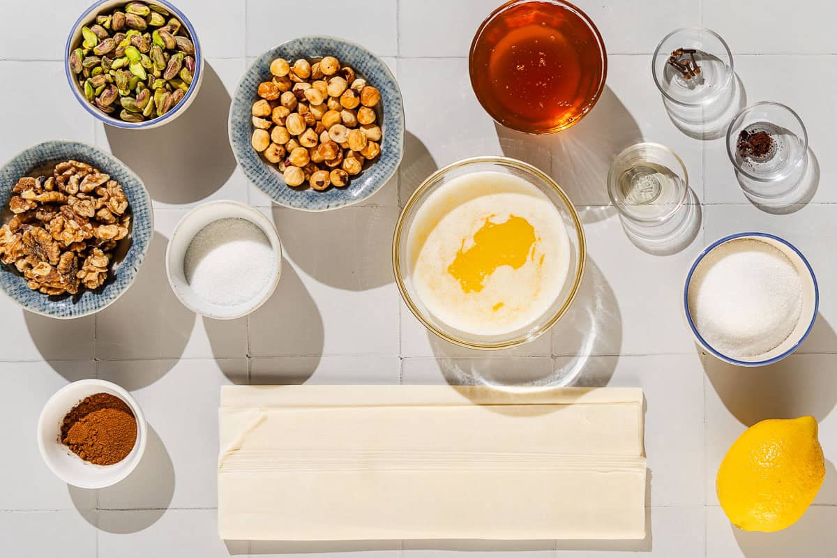 Ingredients for baklava including pistachios, walnuts, hazelnuts, sugar, ground cinnamon, cloves, phyllo dough, butter, sugar, honey, orange extract, and lemon.