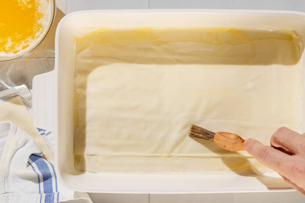 A sheet of phyllo dough being brushed with butter in a baking dish next to a bowl of melted butter.
