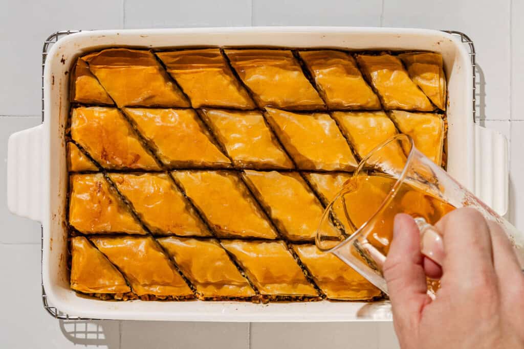 The honey syrup being poured on the sliced baklava in a baking dish.