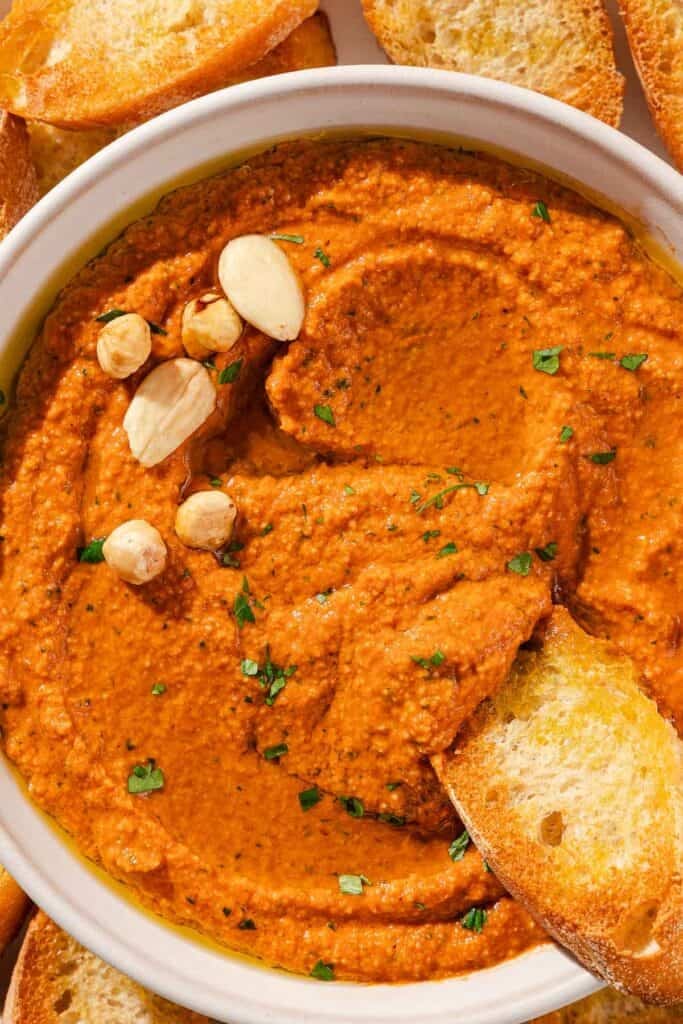 A slice of crusty bread being dipped into a bowl of romesco sauce.