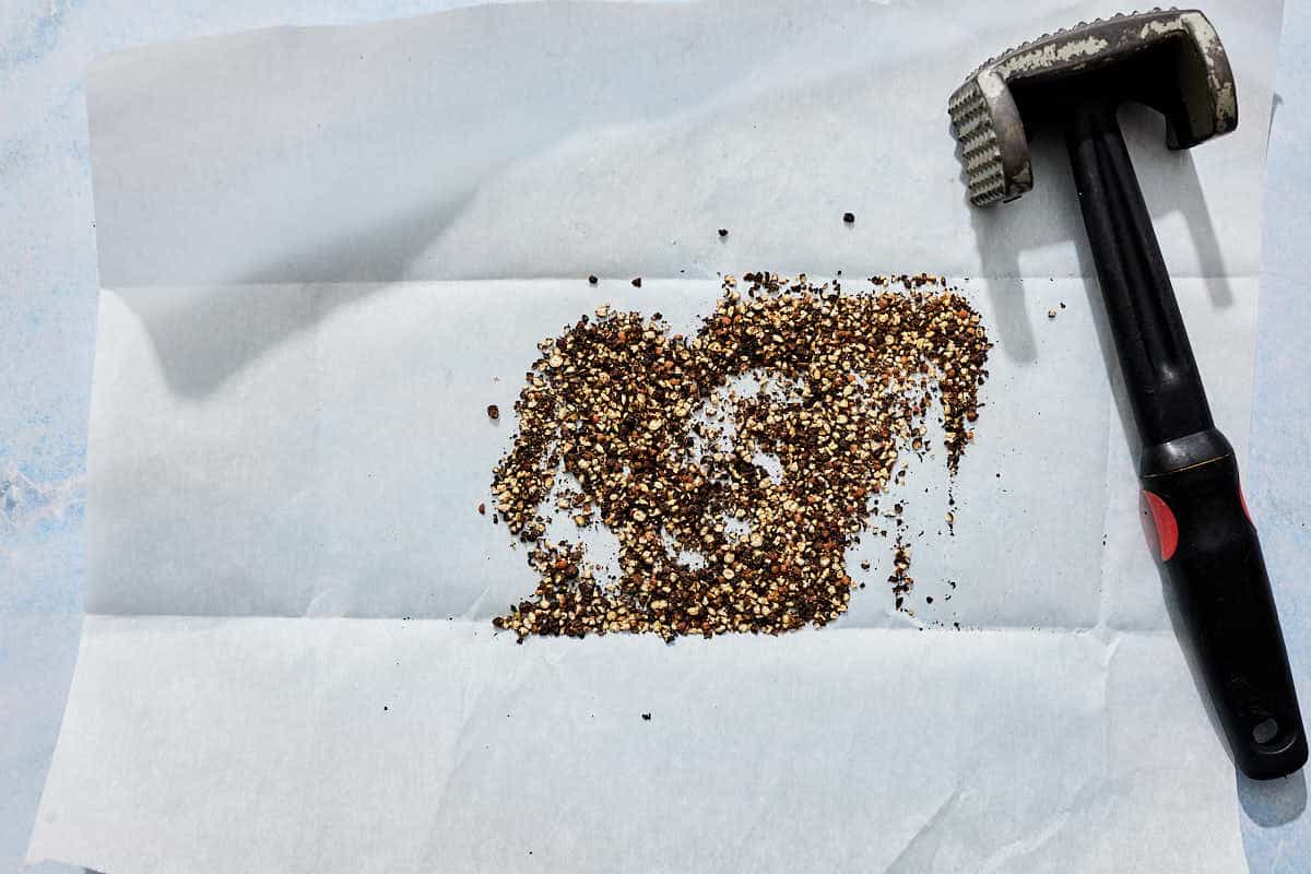 cracked black peppercorns next to a mallet on a piece of parchment paper.