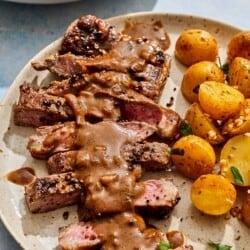 A serving of steak au poivre topped with sauce on a plate with potatoes.