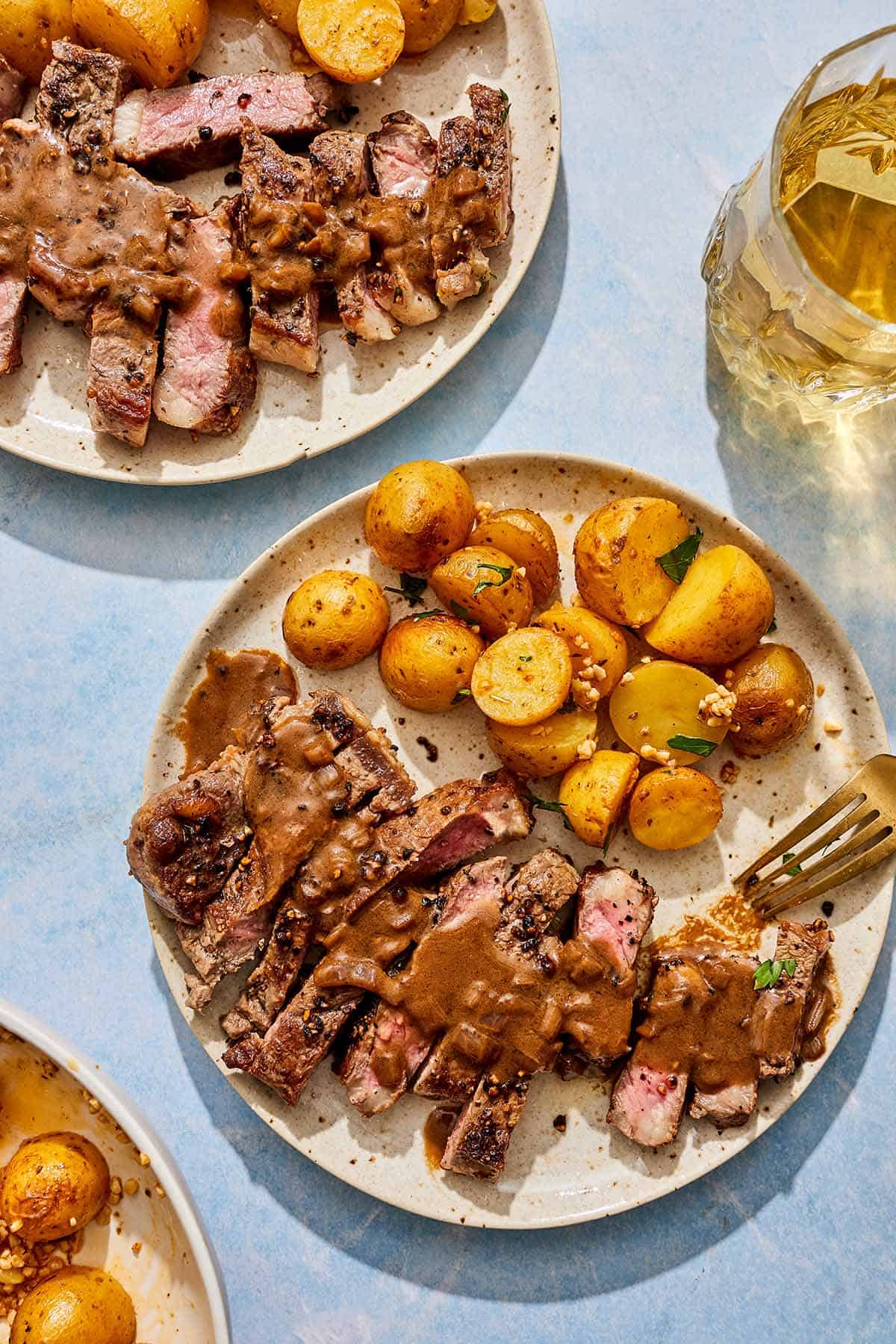 Two servings of steak au poivre topped with sauce on a plate with potatoes next to a glass of wine.