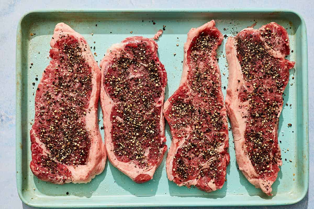 Uncooked steaks for the steak au poivre with a peppercorn crust on a sheet pan.
