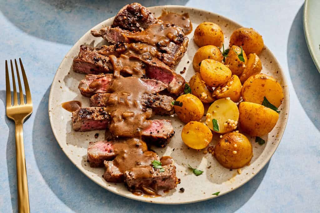 A serving of steak au poivre topped with sauce on a plate with potatoes.