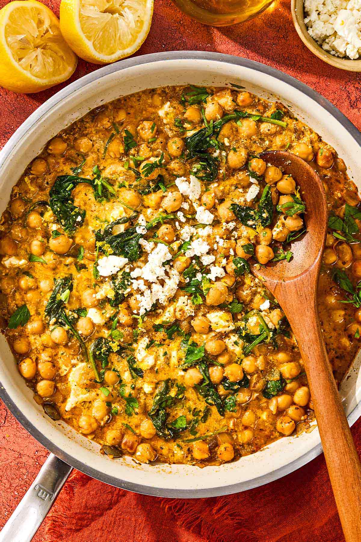 A chickpea skillet with a wooden spoon next to two juiced lemon halves and a bowl of crumbled feta.