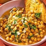 A serving of the chickpea skillet in a bowl with a slice of toast and a fork. Next to this is a bowl of crumbled feta.