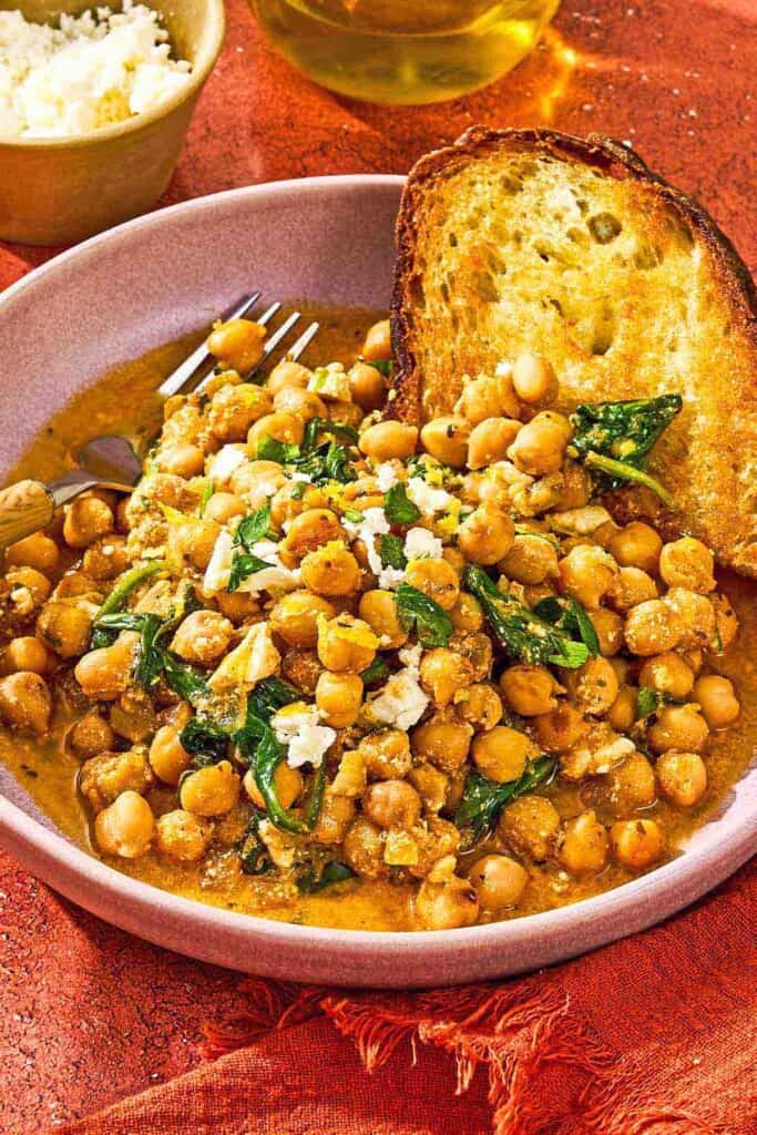A serving of the chickpea skillet in a bowl with a slice of toast and a fork. Next to this is a bowl of crumbled feta.