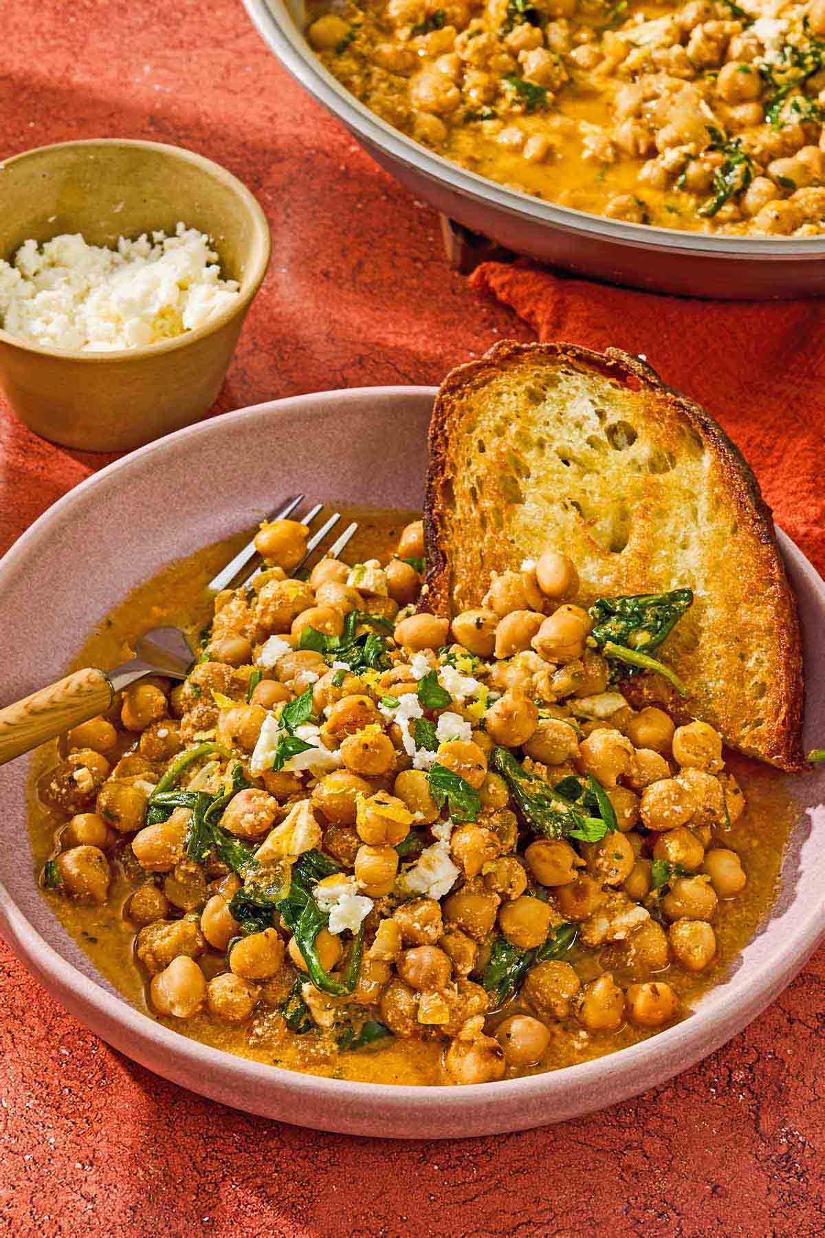 A serving of the chickpea skillet in a bowl with a slice of toast and a fork. Next to this is a bowl of crumbled feta.