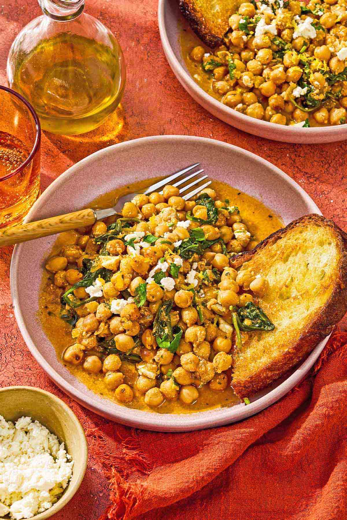 A serving of the chickpea skillet in a bowl with a slice of toast and a fork. Next to this is another serving of the recipe in a bowl, a bowl of crumbled feta, a bottle of olive oil, and a glass of sparkling water.