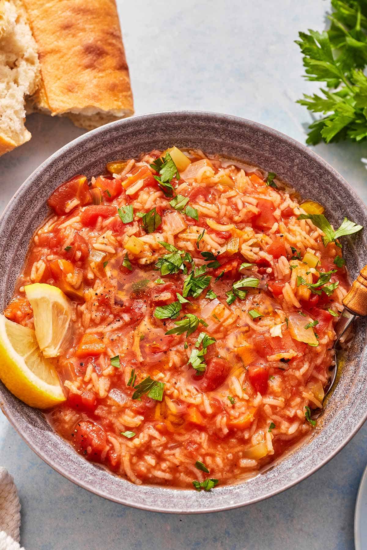 Tomato rice soup in a bowl with lemon wedges and a spoon next to some crusty bread and parsley.