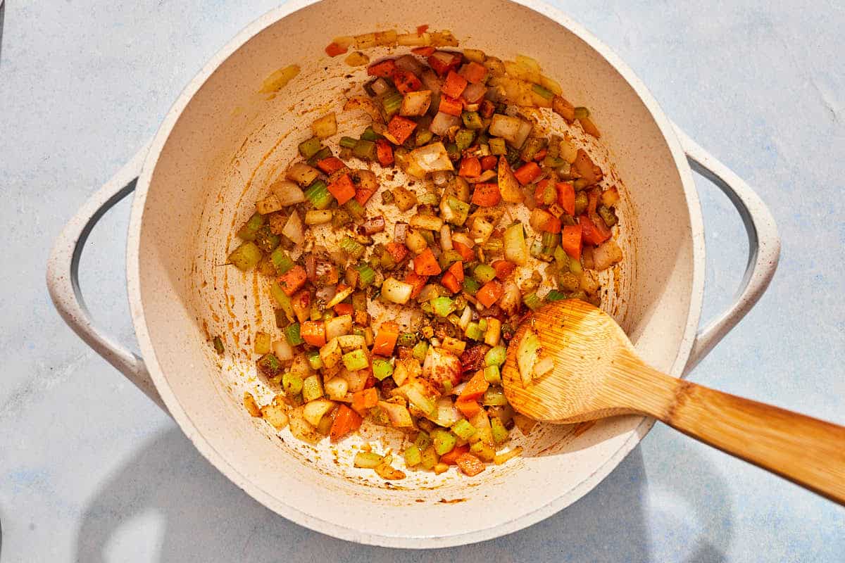 The the vegetables for the tomato rice soup being sautéed with the spices in a pot with a wooden spoon.