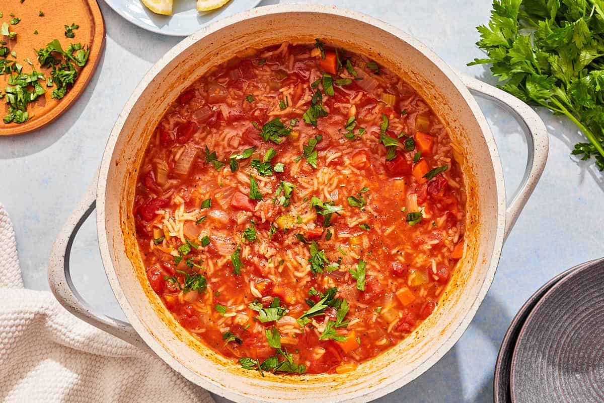 Tomato rice soup garnished with parsley in a large pot next to a stack of two bowls and parsley.